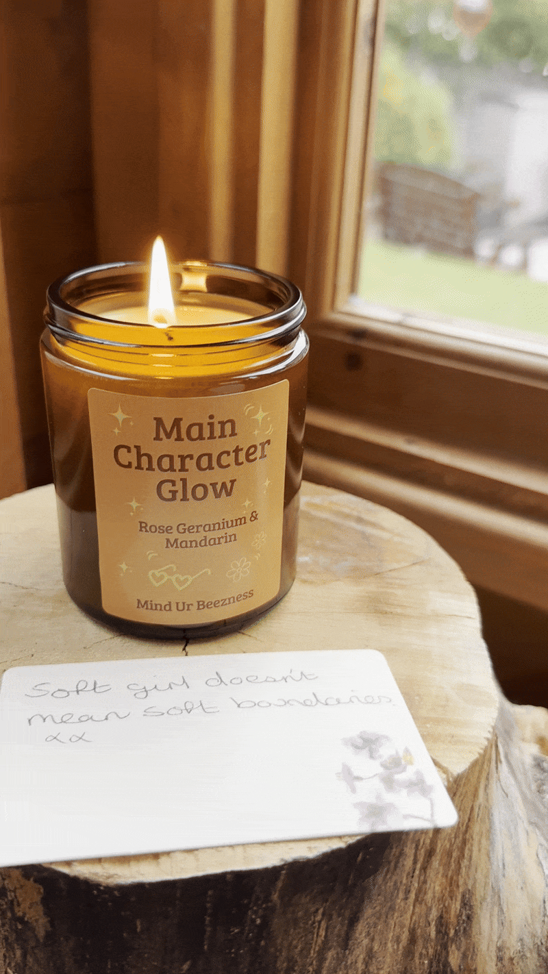 Main Character Glow candle by Mind Ur Beezness, burning on a wooden surface next to a handwritten note that reads “Soft girl doesn’t mean soft boundaries.”
