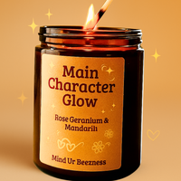 Main Character Glow candle by Mind Ur Beezness, lit with a match in a warm orange soft girl aesthetic, featuring rose geranium and mandarin essential oils.
