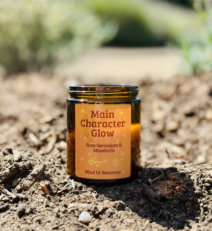 Main Character Glow candle by Mind Ur Beezness placed in earthy soil outdoors, amber jar with orange label — rose geranium and mandarin scent for grounding self-care rituals in nature
