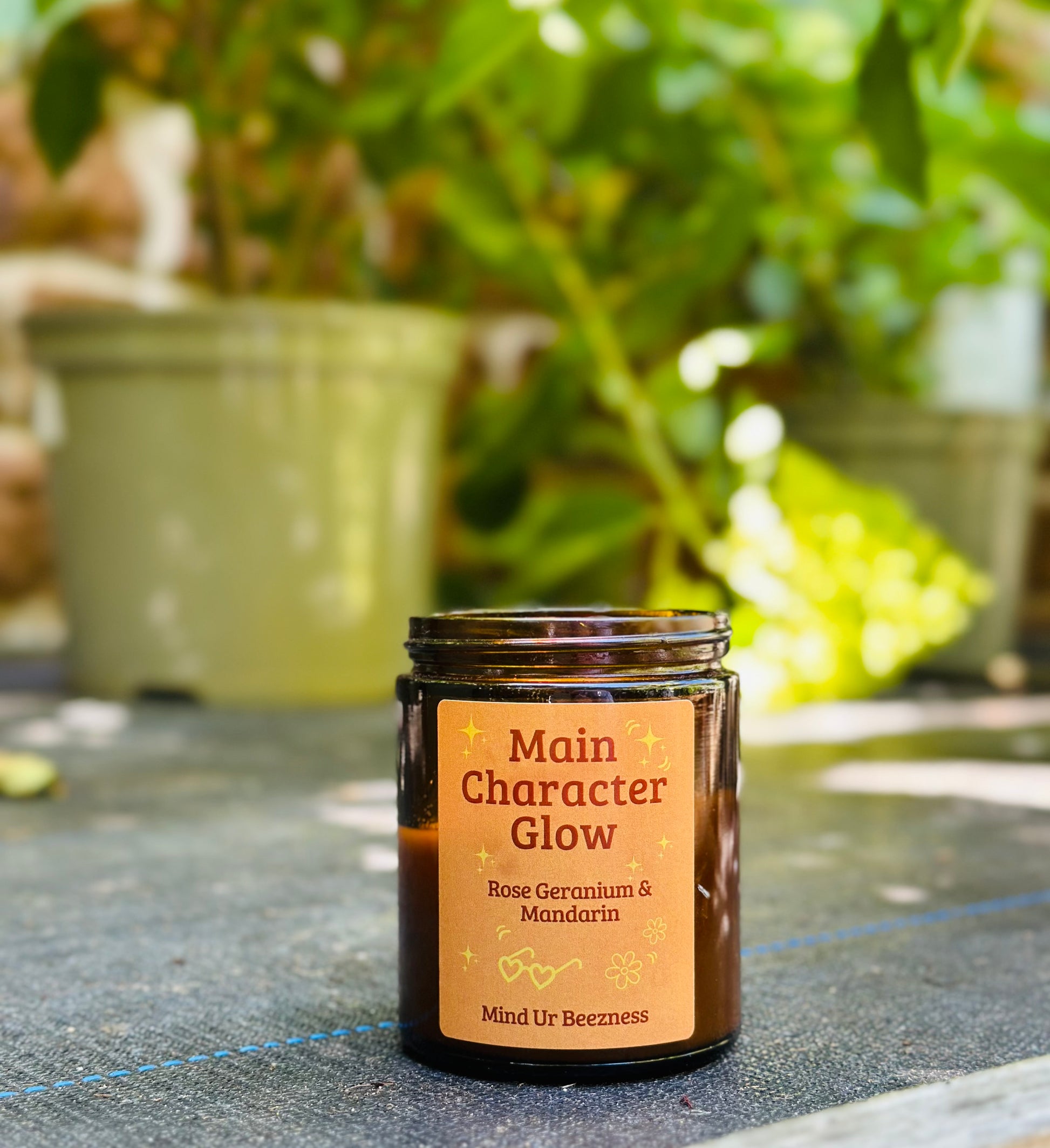 Amber jar Main Character Glow candle by Mind Ur Beezness with rose geranium and mandarin scent, placed on an outdoor table surrounded by green potted plants — soft girl aesthetic for mindful mornings
