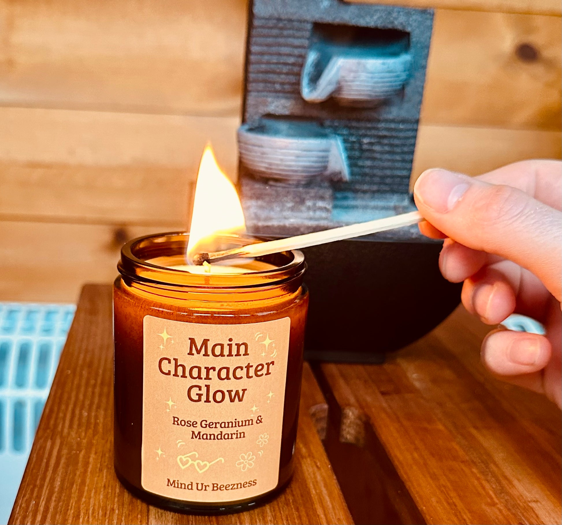 Main Character Glow candle by Mind Ur Beezness being lit with a matchstick in a warm, wooden setting next to a mini tabletop fountain — a soft girl evening ritual.