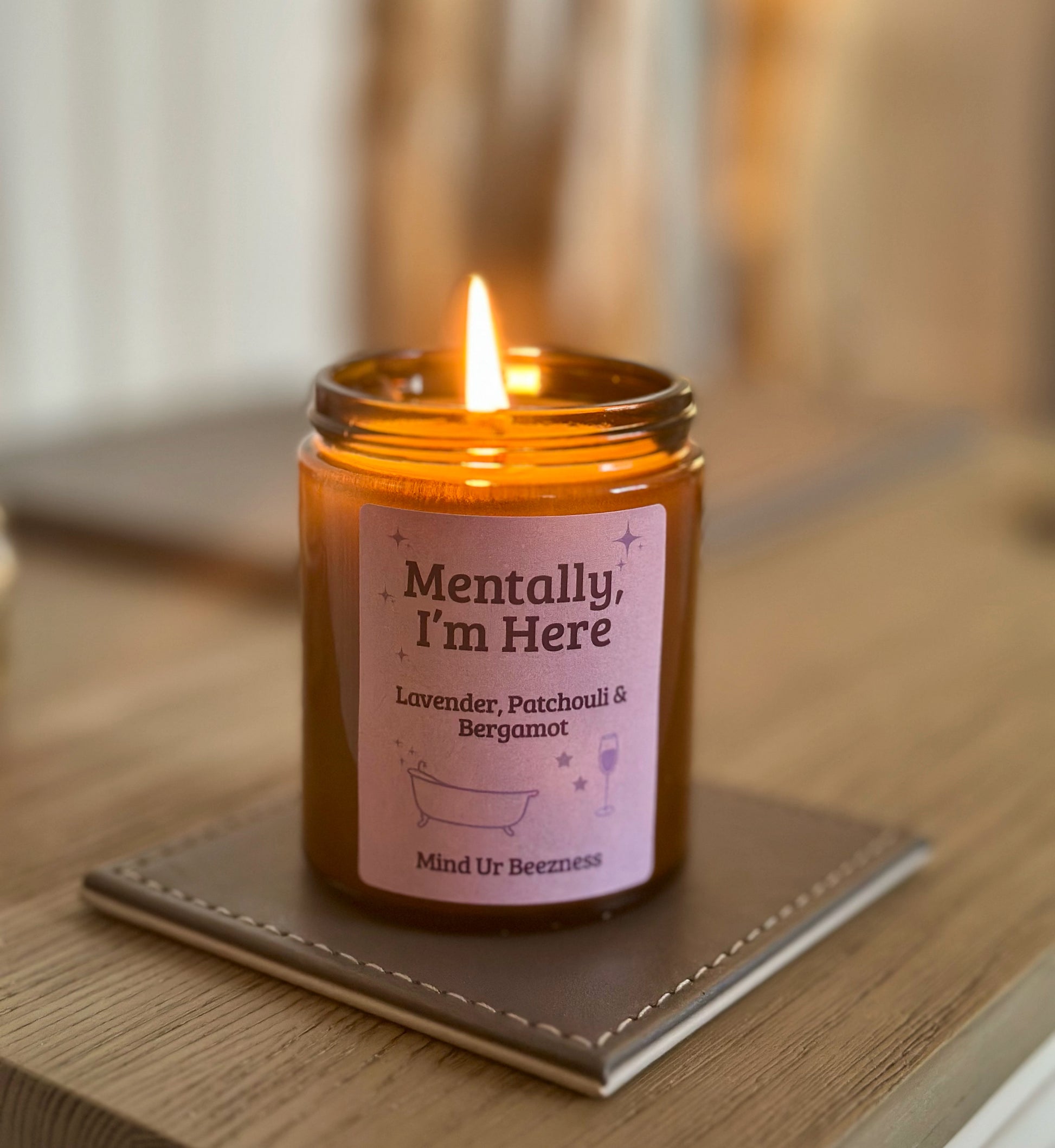 Amber glass candle labeled “Mentally, I’m Here” burning on a wooden table, featuring lavender, patchouli, and bergamot scent by Mind Ur Beezness.

