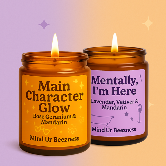 Two amber glass candles from Mind Ur Beezness — Main Character Glow with Rose Geranium & Mandarin, and Mentally, I’m Here with Lavender, Vetiver & Mandarin — set against a soft purple and orange backdrop.
