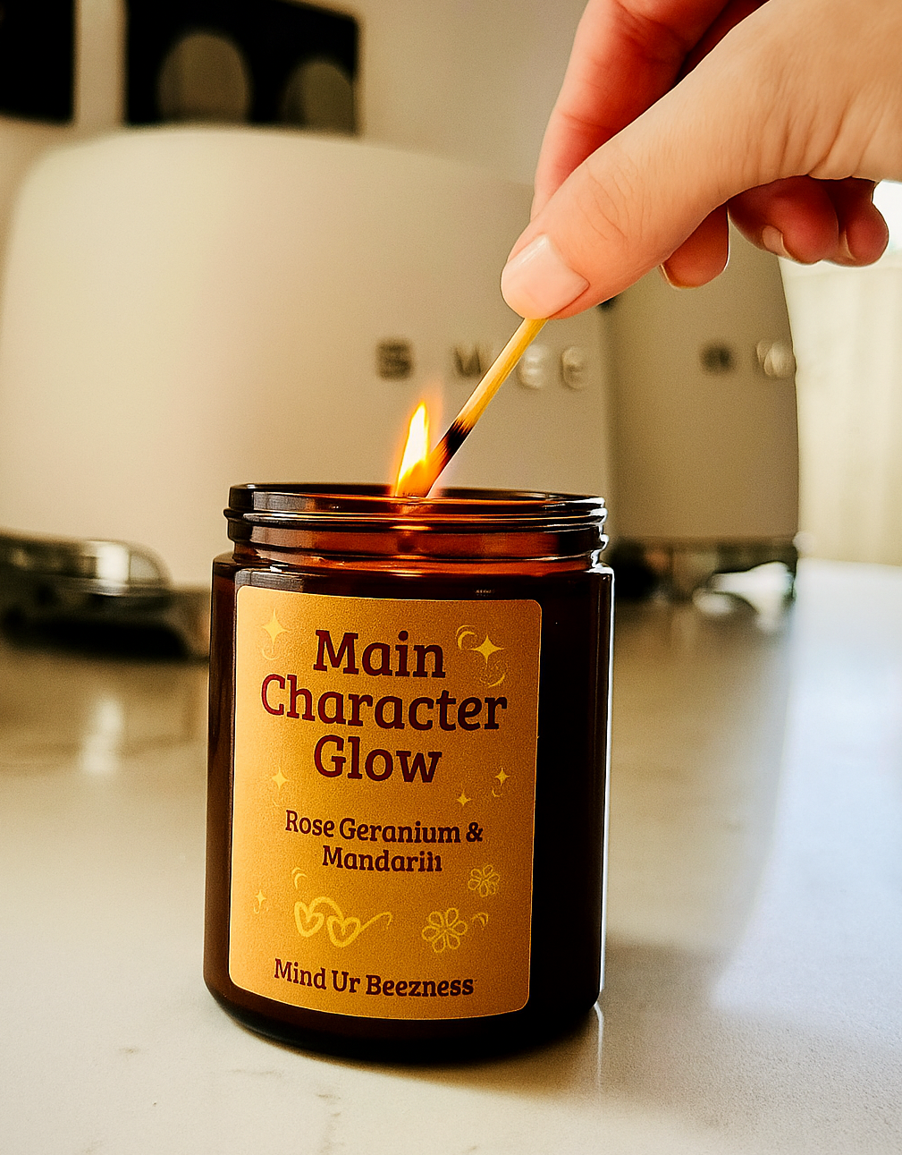 Hand lighting the “Main Character Glow” candle by Mind Ur Beezness with a matchstick. Candle sits on a clean kitchen surface, with a blurred SMEG appliance in the background.

