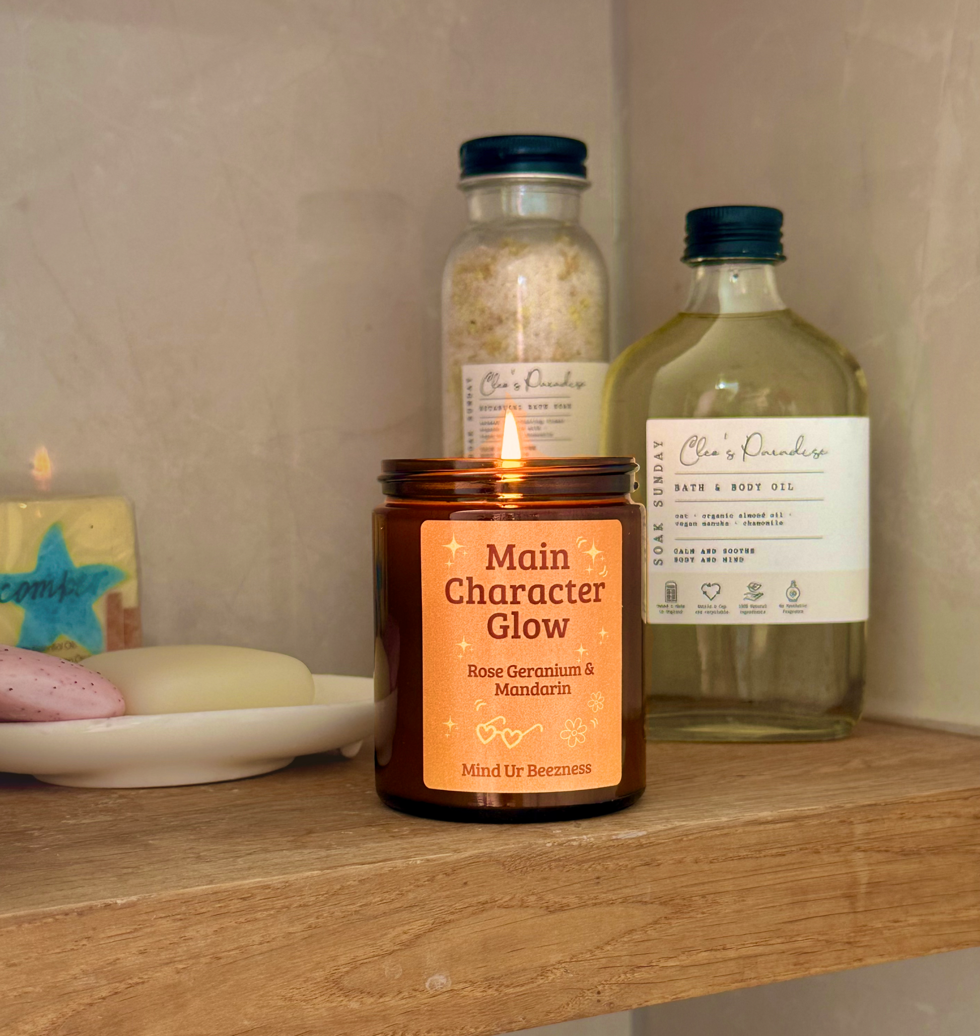 Lit “Main Character Glow” candle on a wooden bathroom shelf beside bath oil, salts, and soap bars. Cozy self-care vibe with neutral stone tile background.
