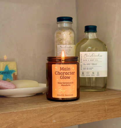 Lit “Main Character Glow” candle on a wooden bathroom shelf beside bath oil, salts, and soap bars. Cozy self-care vibe with neutral stone tile background.
