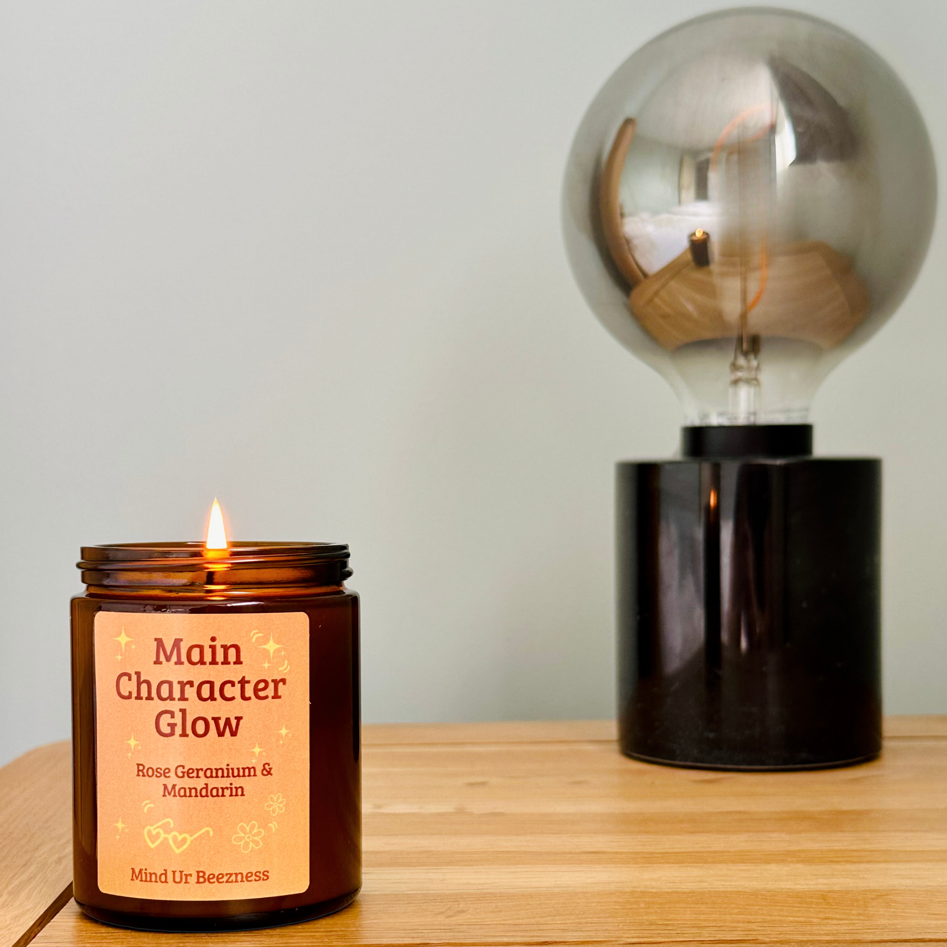 Amber glass “Main Character Glow” candle lit on a wooden surface next to a modern round bulb lamp. Minimalist, calm setting with a neutral wall in the background.
