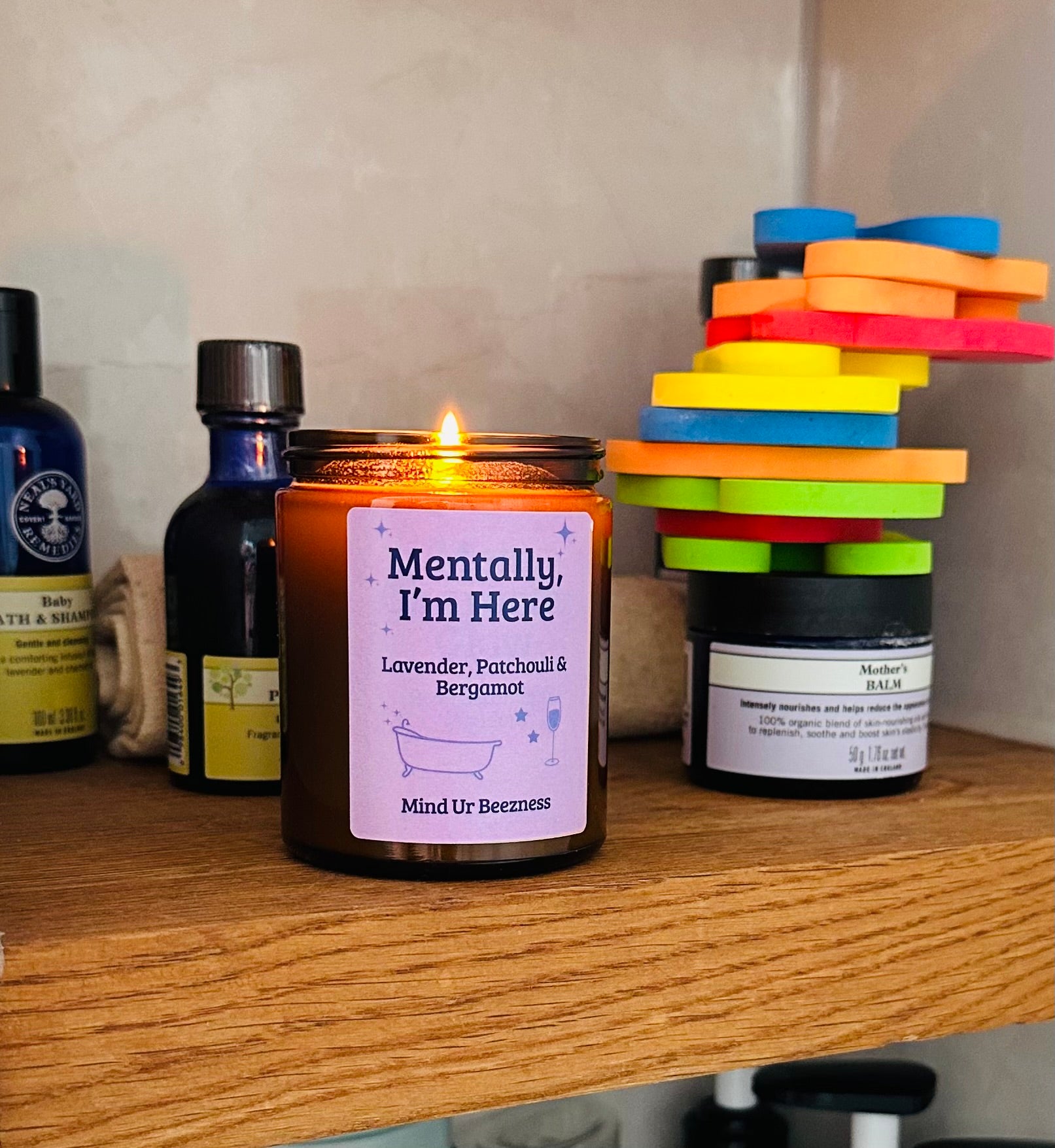 Mind Ur Beezness candle labelled “Mentally, I’m Here” with Lavender, Patchouli & Bergamot, lit on a wooden bathroom shelf surrounded by skincare products and colourful bath toys.
