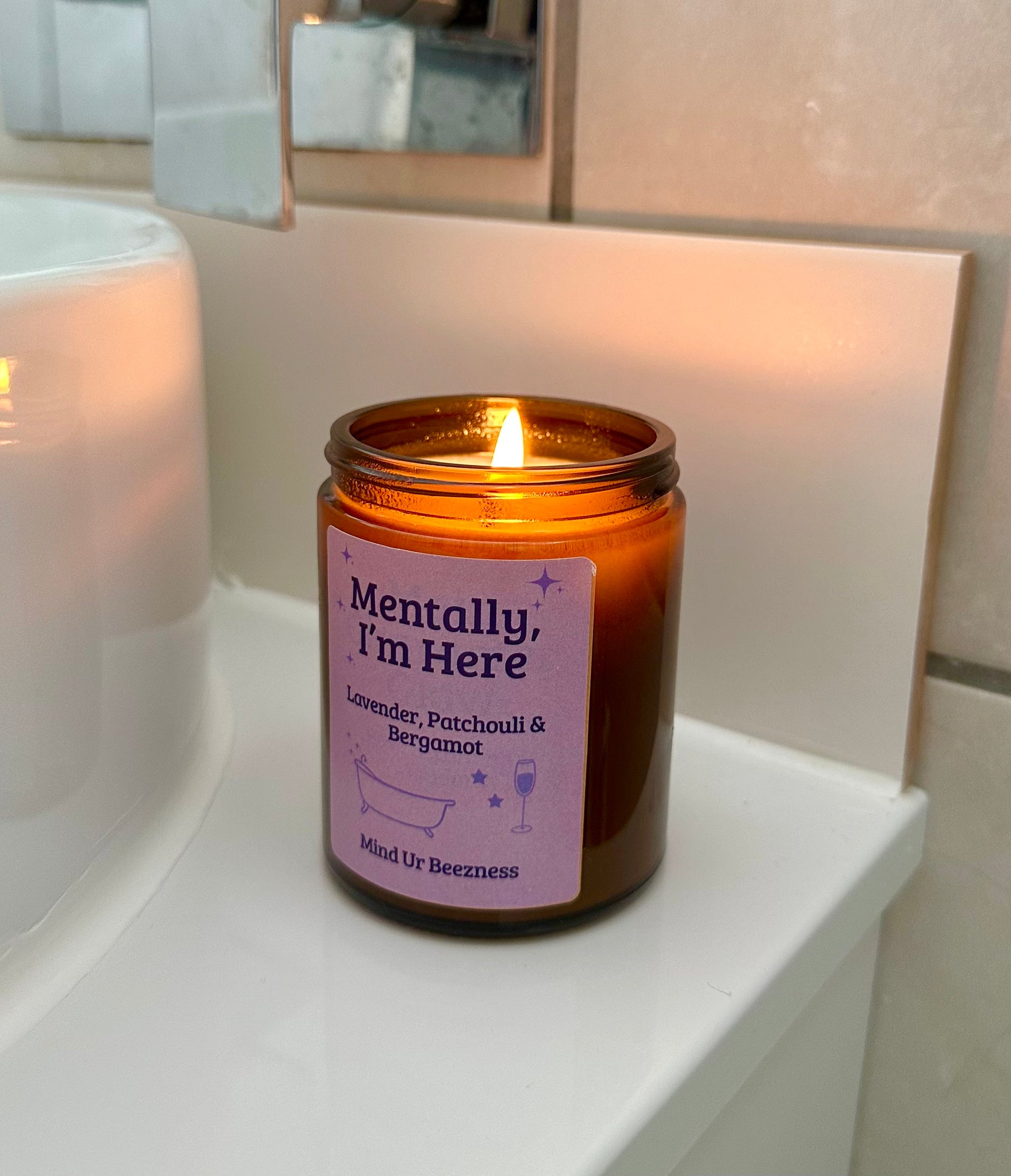 Mind Ur Beezness “Mentally, I’m Here” candle with Lavender, Patchouli & Bergamot, softly glowing next to a modern bathroom sink and chrome faucet.
