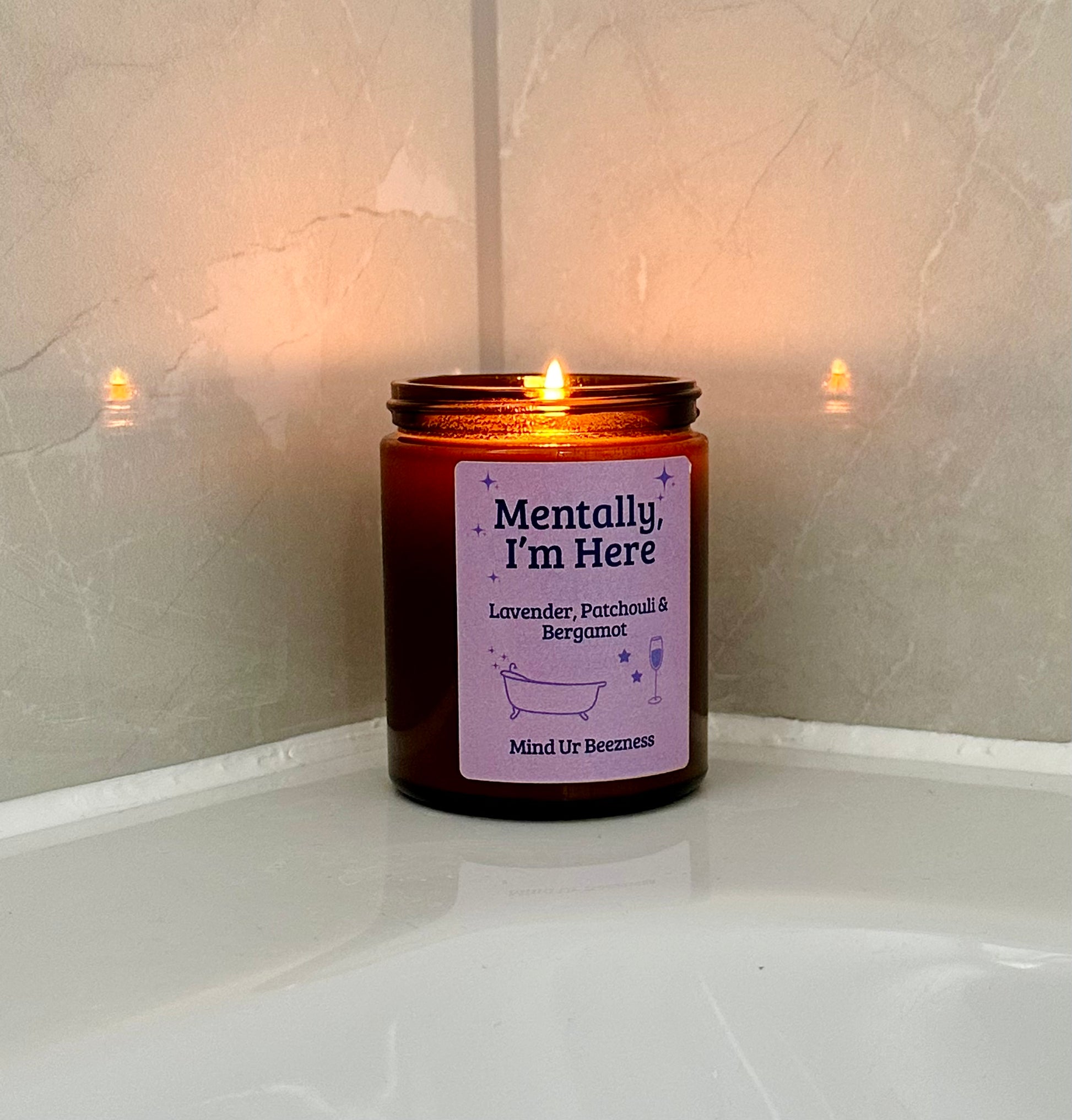 Mind Ur Beezness “Mentally, I’m Here” candle with Lavender, Patchouli & Bergamot, lit beside a white bathtub, casting a soft reflection on glossy bathroom tiles.