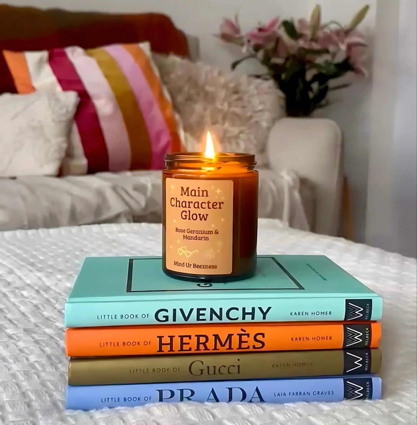 A lit “Main Character Glow” soft girl era candle by Mind Ur Beezness, styled on a stack of designer fashion books in a cozy, feminine bedroom.

